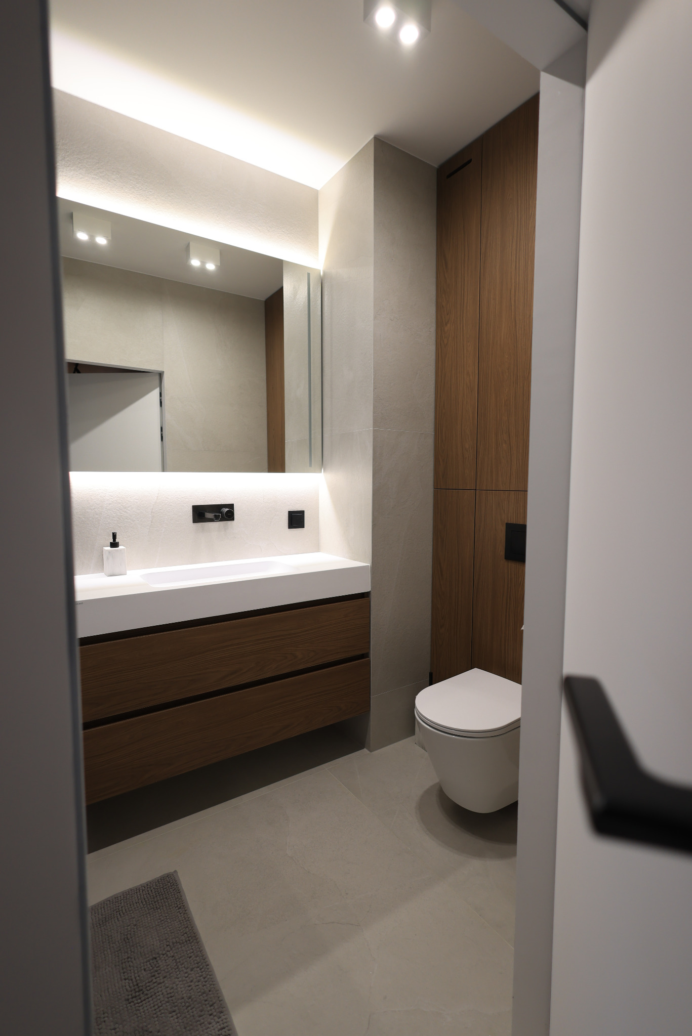 Nowoczesna łazienka z drewnianą szafką pod umywalką, podświetlanym lustrem i podwieszaną toaletą, widziana zza uchylonych drzwi.