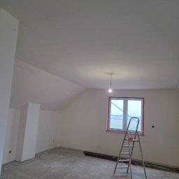 KZW INVEST - Pokój na poddaszu po szpachlowaniu ścian i sufitu, z widoczną drabiną malarską i oknem w czerwonej ramie, przygotowany do dalszych prac wykończeniowych.