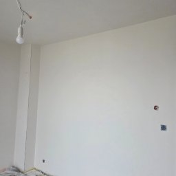 KZW INVEST - Wnętrze po malowaniu na biało, widoczne gniazdka i zabezpieczona podłoga folią malarską. Wisząca żarówka na kablu. Świeżo pomalowane ściany i sufit.