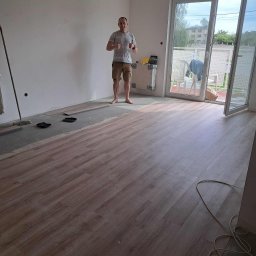 KZW INVEST - Połowa pokoju z ułożonymi jasnymi panelami, druga w trakcie układania. Mężczyzna w tle pokazuje kciuki w górę. Widok na balkon i okno.