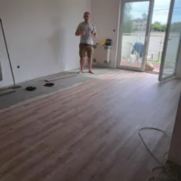 Połowa pokoju z ułożonymi jasnymi panelami, druga w trakcie układania. Mężczyzna w tle pokazuje kciuki w g&oacute;rę. Widok na balkon i okno.