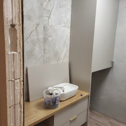 KZW INVEST - Remont łazienki w toku: odsłonięta ściana z cegły, marmurowe płytki, biała umywalka na szafce z drewnianym blatem, wisząca szafka. Widoczne narzędzia i akcesoria.