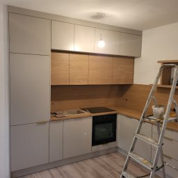 KZW INVEST - Wnętrze kuchni w trakcie montażu: szare szafki z drewnianymi akcentami, piekarnik i drabina malarska. Podłoga częściowo pokryta panelami.