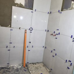 KZW INVEST - Ściany w trakcie układania jasnych, kwadratowych płytek ceramicznych. Widoczny poziomica, krzyżyki dystansowe i fragment surowej ściany z zaprawą.
