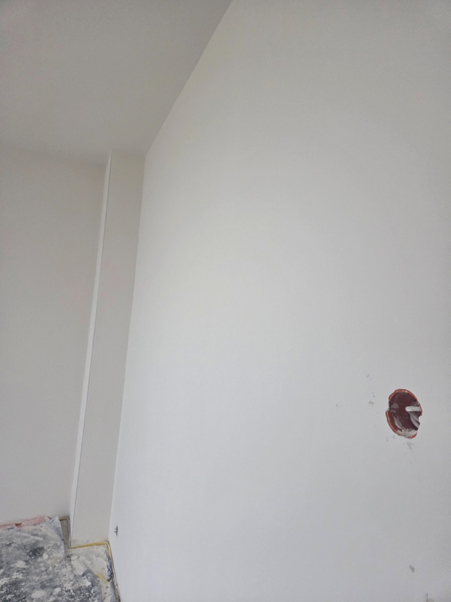 Ściana po szpachlowaniu i malowaniu na biało, widoczna puszka elektryczna i fragment podłogi z folią ochronną. Minimalistyczny kadr, świeżo wykończone wnętrze.