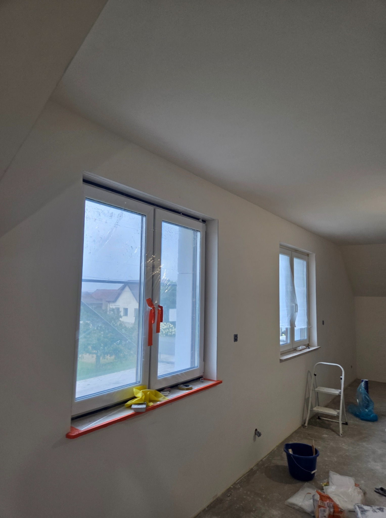 Pokój na poddaszu po szpachlowaniu ścian, przygotowany do malowania. Okna zabezpieczone folią, widoczna drabina i wiadro.