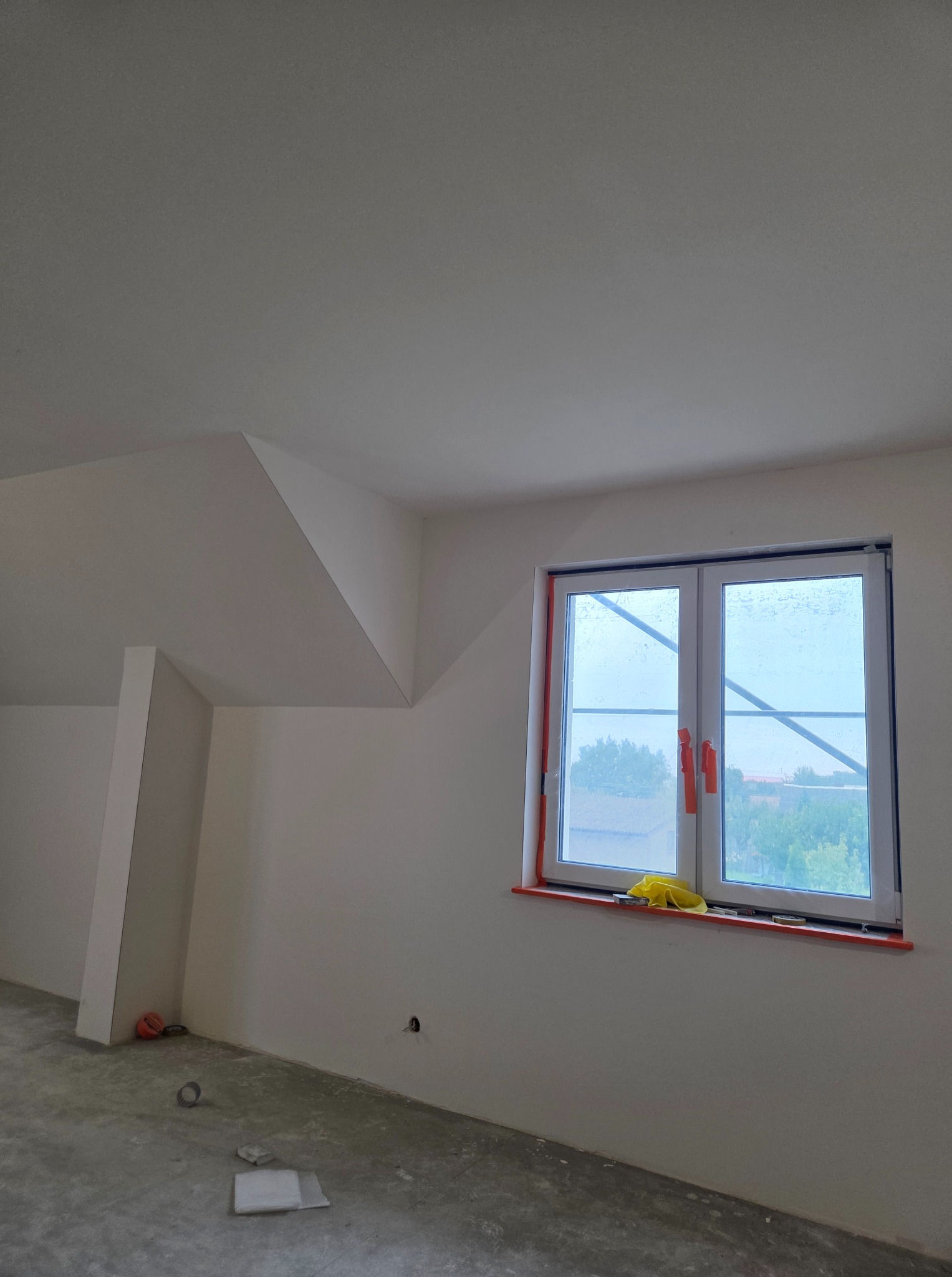Wnętrze poddasza po szpachlowaniu i malowaniu ścian na biało, widoczne okno z zabezpieczeniami, surowa podłoga i narzędzia.