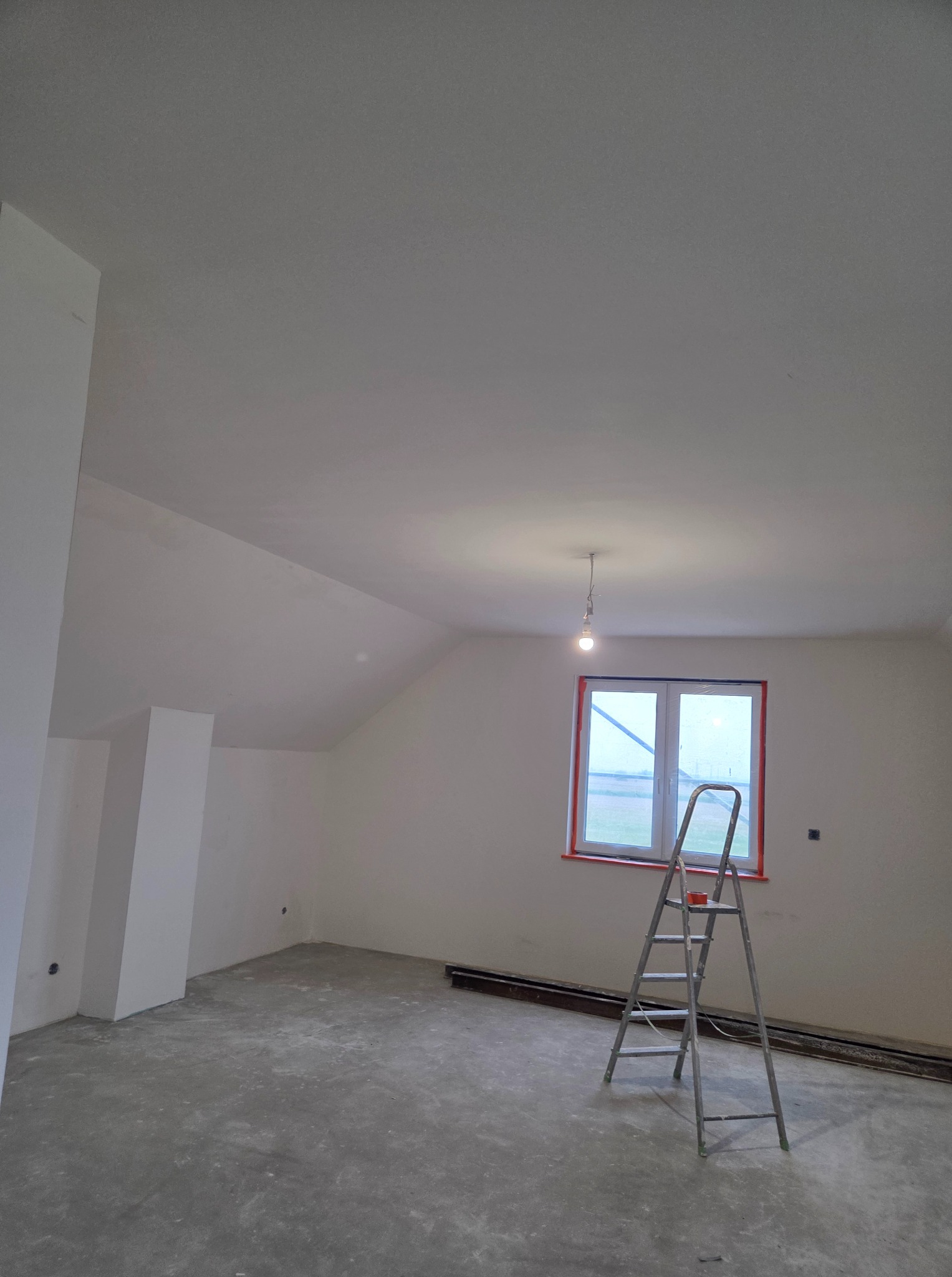 Pokój na poddaszu po szpachlowaniu ścian i sufitu, z widoczną drabiną malarską i oknem w czerwonej ramie, przygotowany do dalszych prac wykończeniowych.