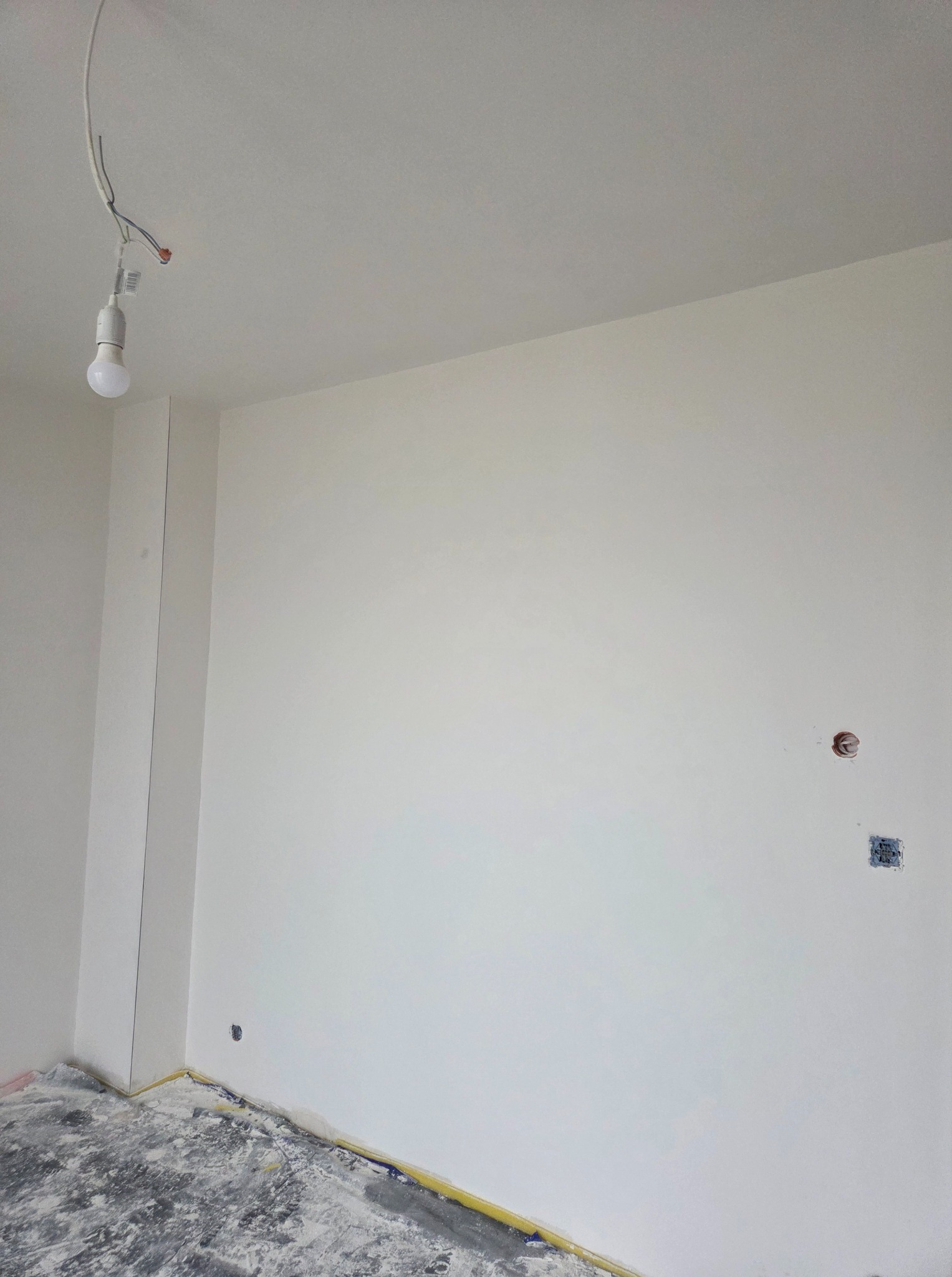 Wnętrze po malowaniu na biało, widoczne gniazdka i zabezpieczona podłoga folią malarską. Wisząca żarówka na kablu. Świeżo pomalowane ściany i sufit.