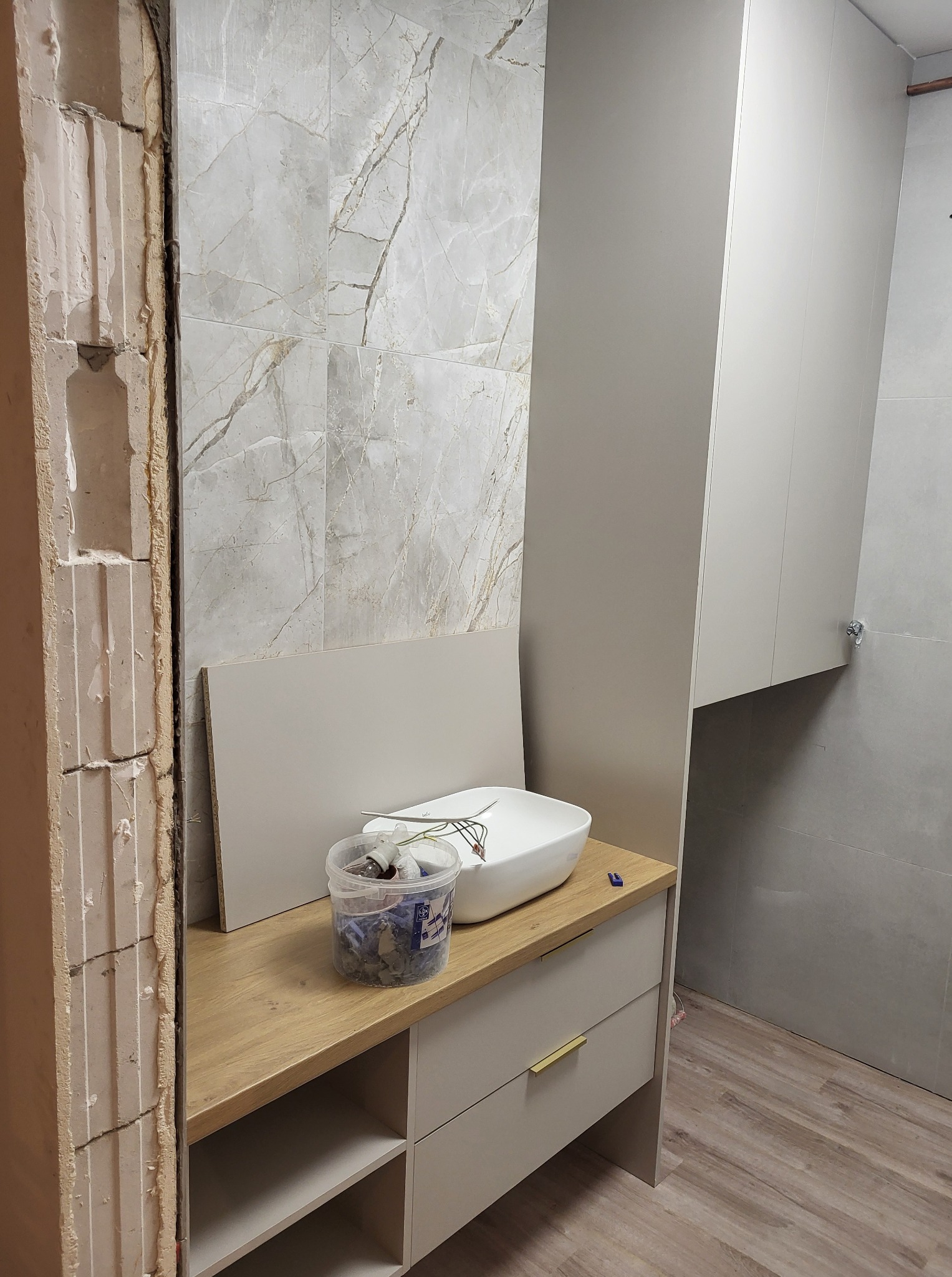 Remont łazienki w toku: odsłonięta ściana z cegły, marmurowe płytki, biała umywalka na szafce z drewnianym blatem, wisząca szafka. Widoczne narzędzia i akcesoria.