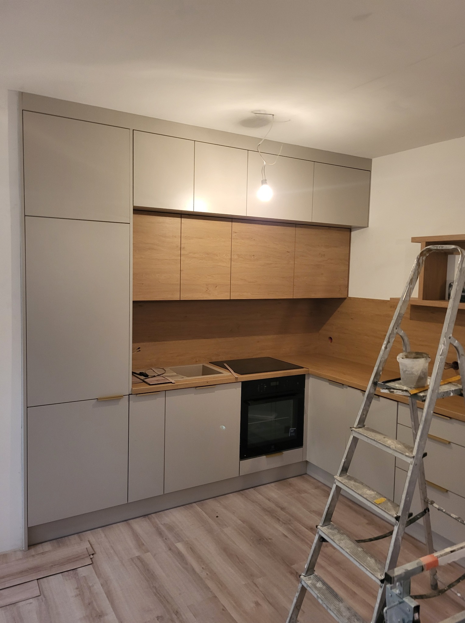 Wnętrze kuchni w trakcie montażu: szare szafki z drewnianymi akcentami, piekarnik i drabina malarska. Podłoga częściowo pokryta panelami.