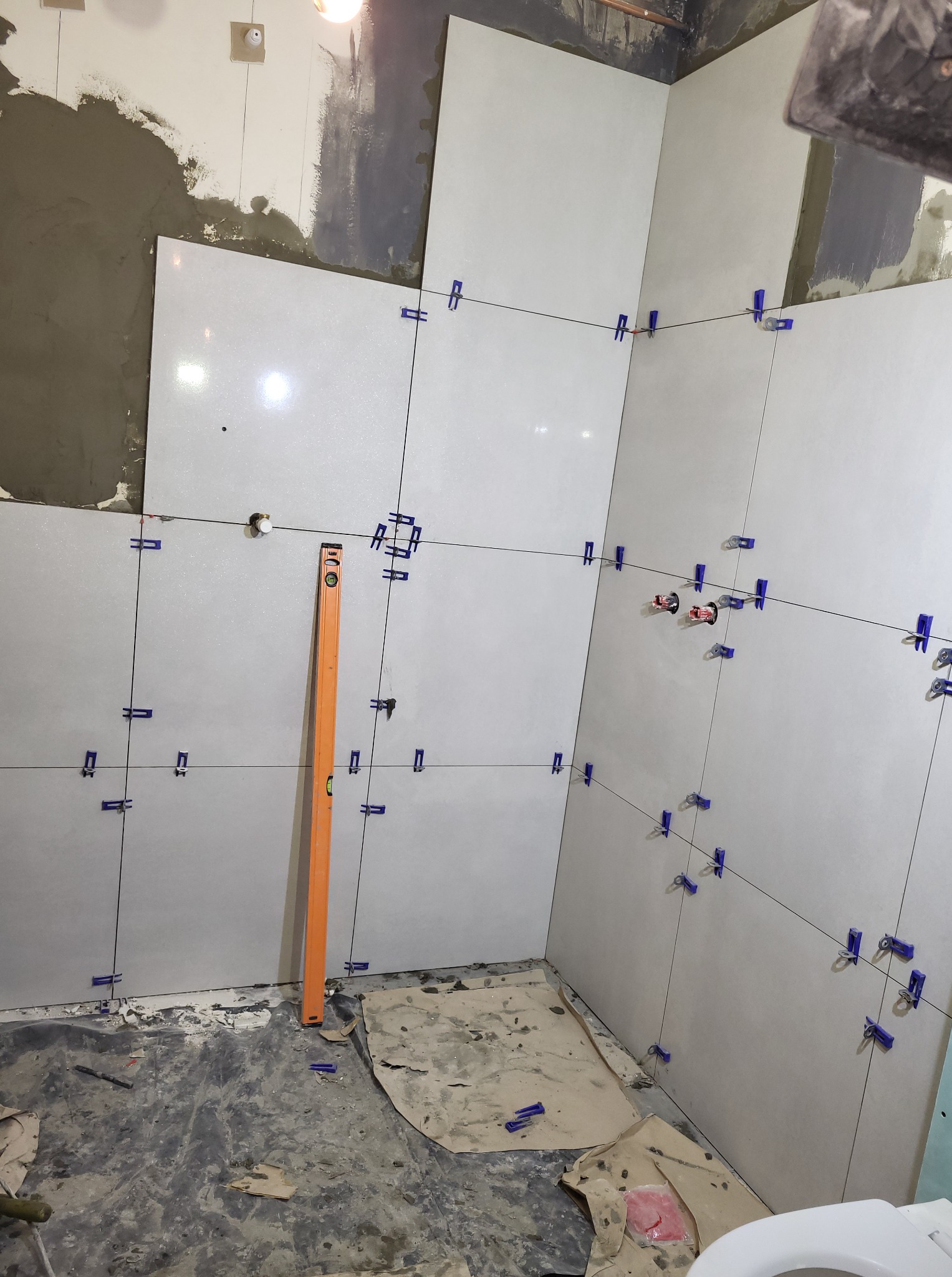 Ściany w trakcie układania jasnych, kwadratowych płytek ceramicznych. Widoczny poziomica, krzyżyki dystansowe i fragment surowej ściany z zaprawą.