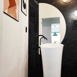 LOLEK Przedsiębiorstwo Handlowo Usługowe Marcin Plewak - Nowoczesna łazienka z białą umywalką wolnostojącą i czarną armaturą na tle marmurowych płytek. W tle widoczne lustro z okrągłym wycięciem i dekoracje ścienne.