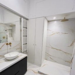 LOLEK Przedsiębiorstwo Handlowo Usługowe Marcin Plewak - Elegancka łazienka po remoncie: białe marmurowe płytki z złotymi żyłami, czarna szafka pod umywalką, złota armatura, kabina prysznicowa typu walk-in i biały grzejnik.
