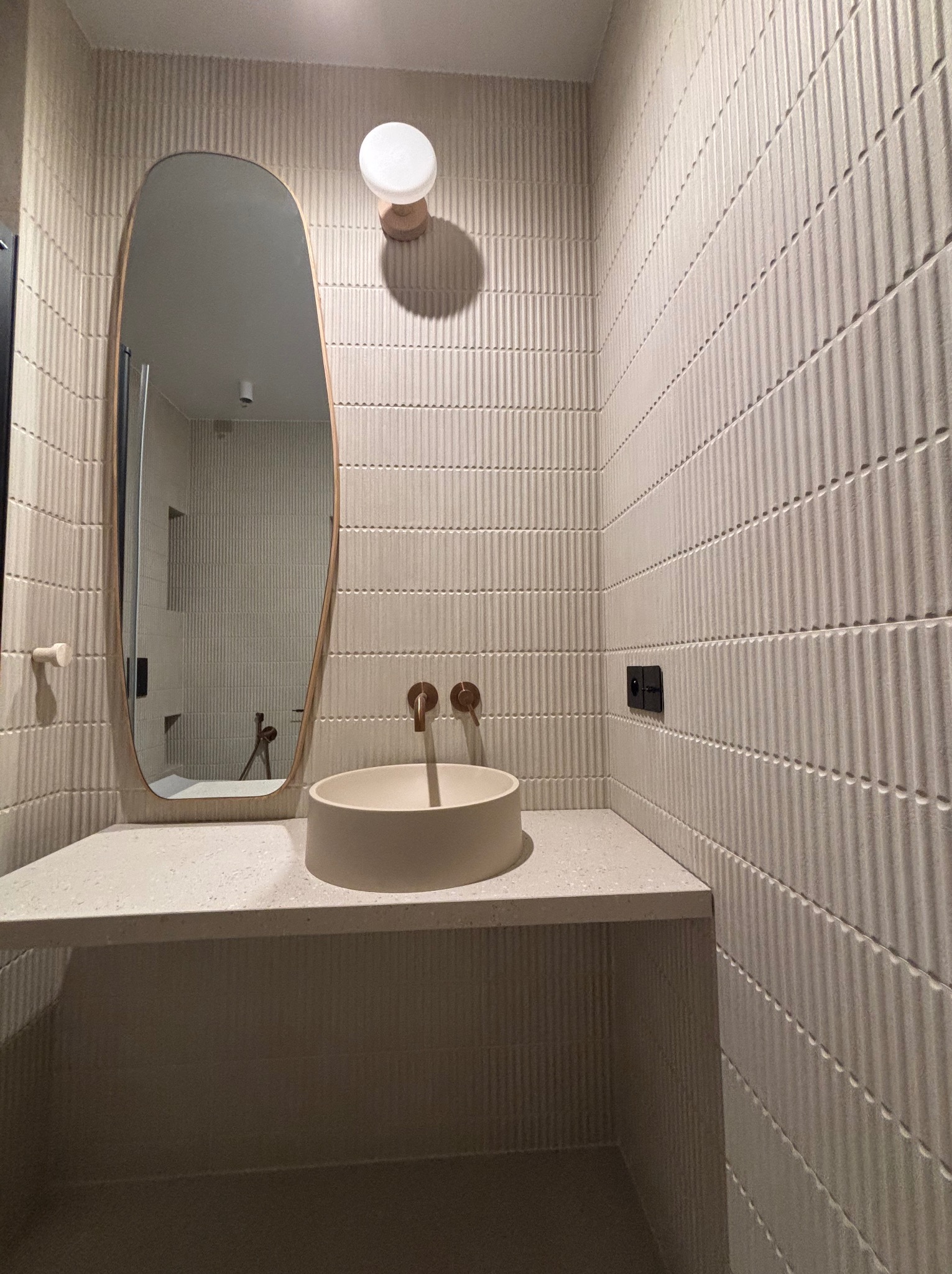 Nowoczesna łazienka z umywalką nablatową, lustrem o organicznym kształcie i teksturowanymi płytkami w odcieniach beżu. Minimalistyczny design, ciepłe oświetlenie.