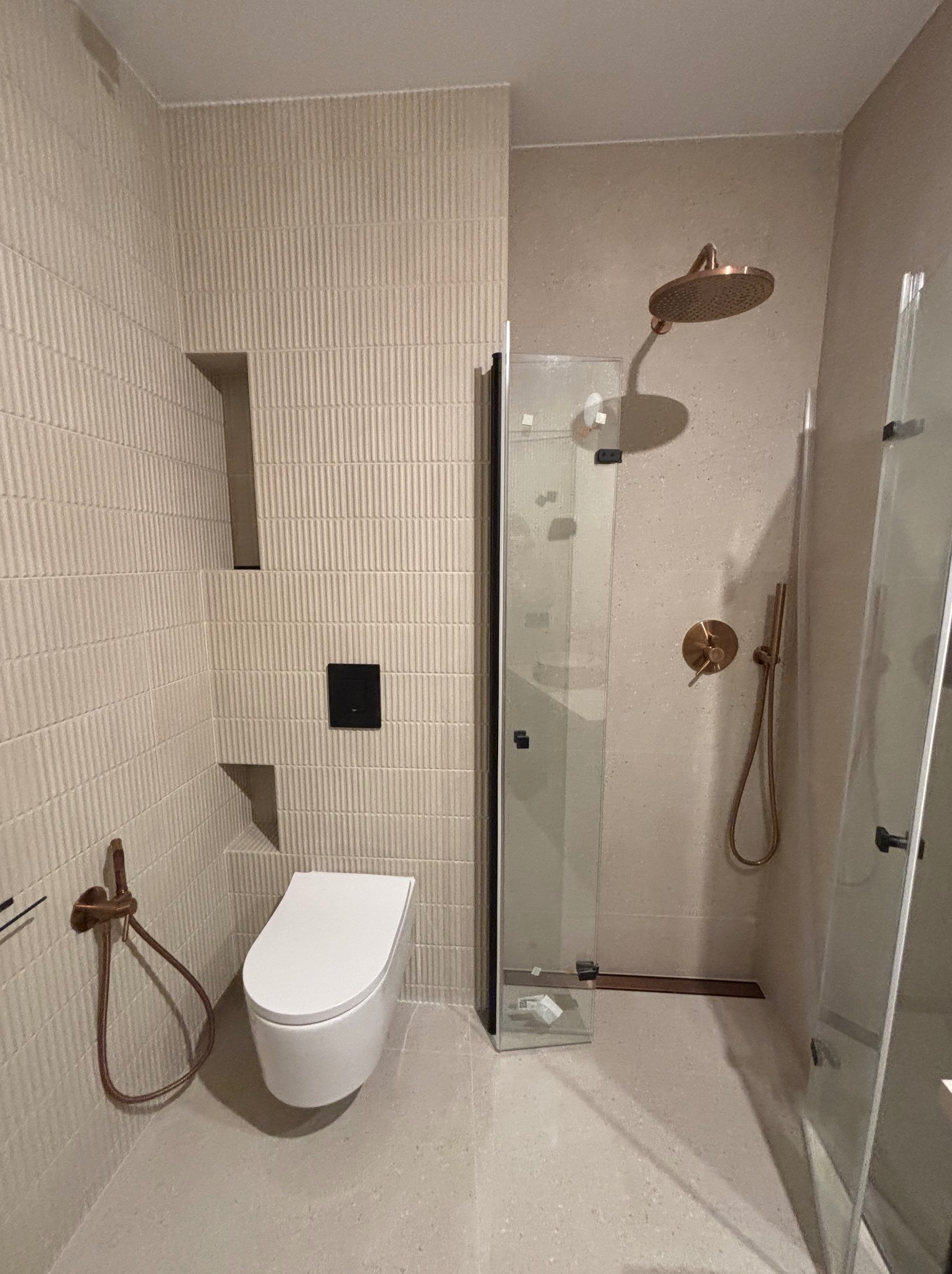 Wykończona łazienka w beżowych odcieniach z deszczownicą w kolorze miedzi, wiszącą toaletą i kabiną prysznicową z czarnymi profilami. Ściany wyłożone płytkami z delikatnym wzorem.