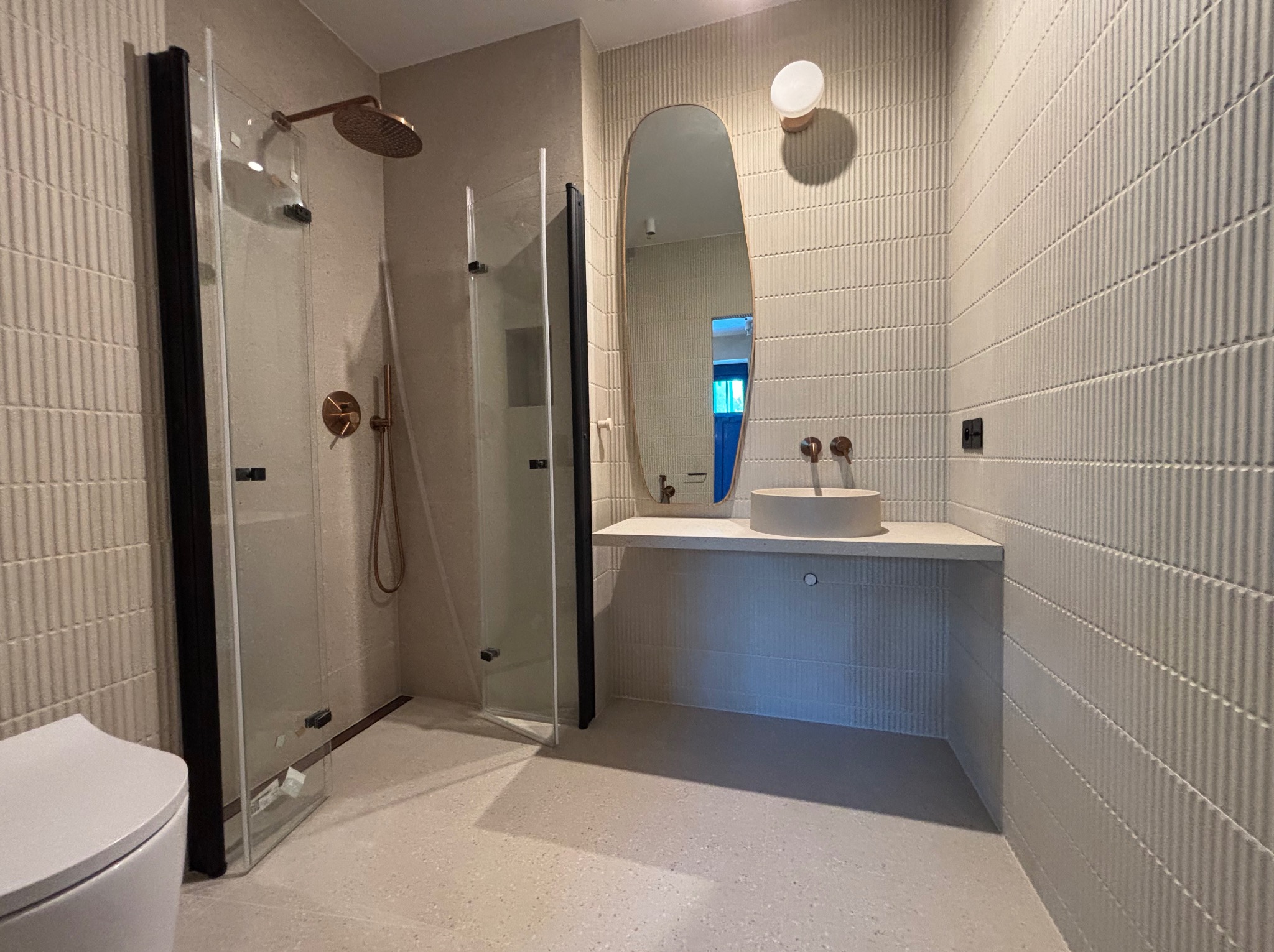 Nowoczesna łazienka z kabiną prysznicową typu walk-in, owalnym lustrem i umywalką nablatową. Beżowe płytki z delikatną fakturą. Armatura w kolorze miedzi.