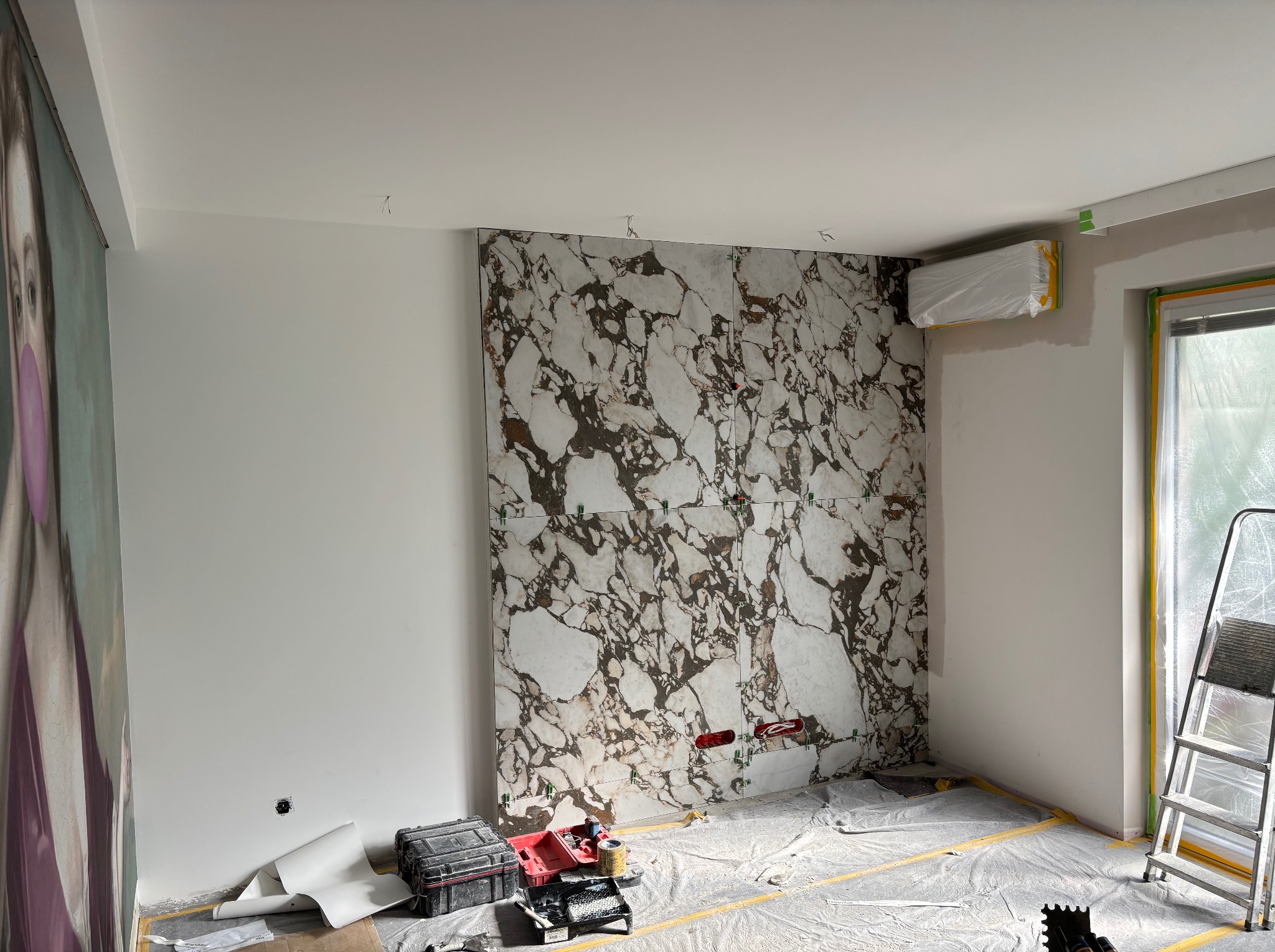 Łazienka w trakcie prac: ściana z płytek imitujących marmur, narzędzia malarskie, drabina. Widoczne zabezpieczenia folią malarską i wystający fragment muralu.