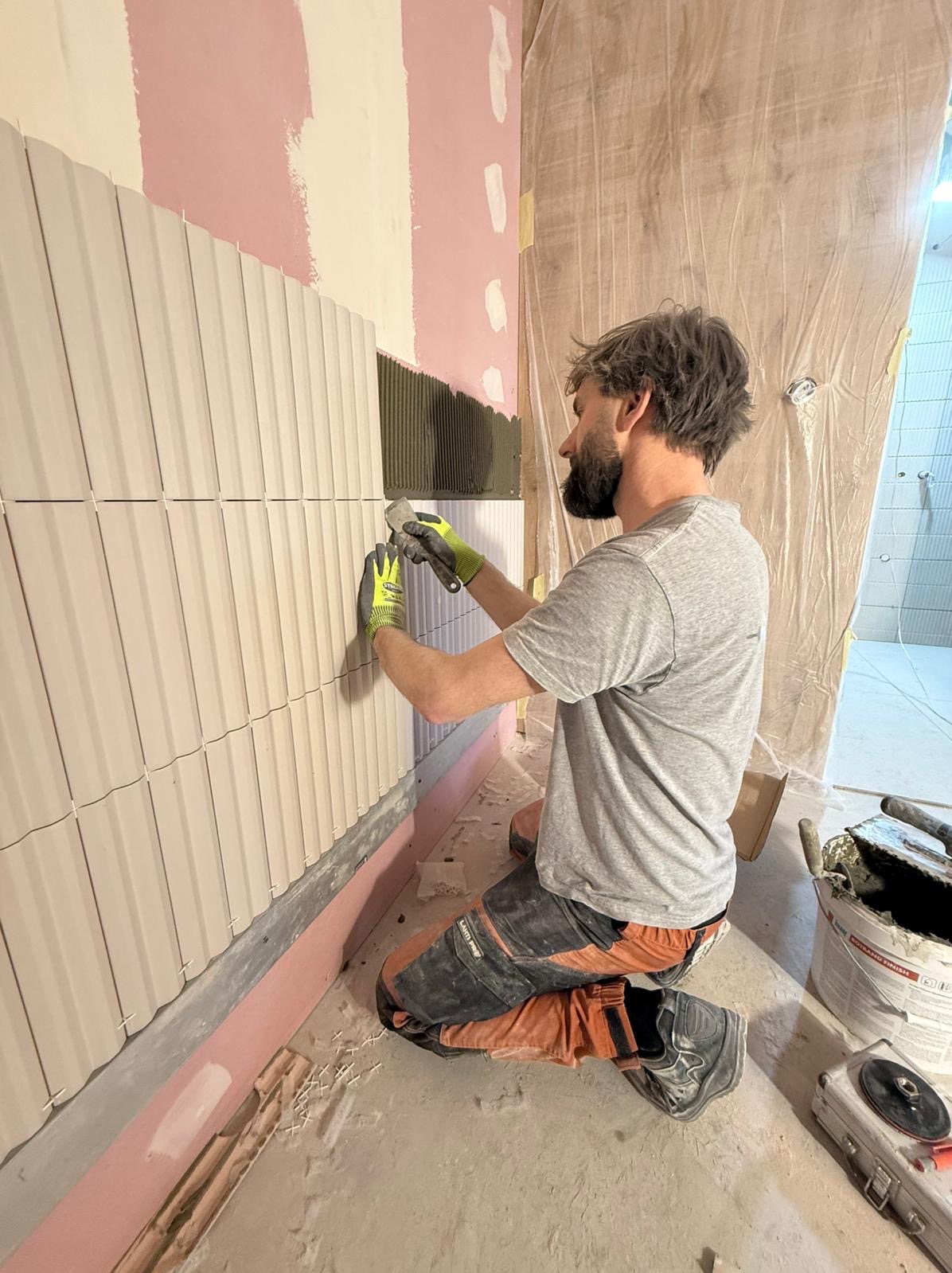 Układanie beżowych, podłużnych płytek na ścianie w łazience przez glazurnika w szarym t-shircie. Widoczne narzędzia i wiadro z klejem. Częściowo zakryta folią ściana.
