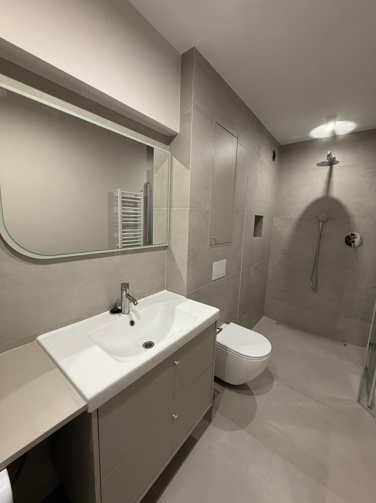 Wykończona łazienka w szarościach: umywalka z szafką, toaleta podtynkowa, kabina prysznicowa typu walk-in. Minimalistyczny design, płytki wielkoformatowe, lustro z zaokrąglonymi rogami.