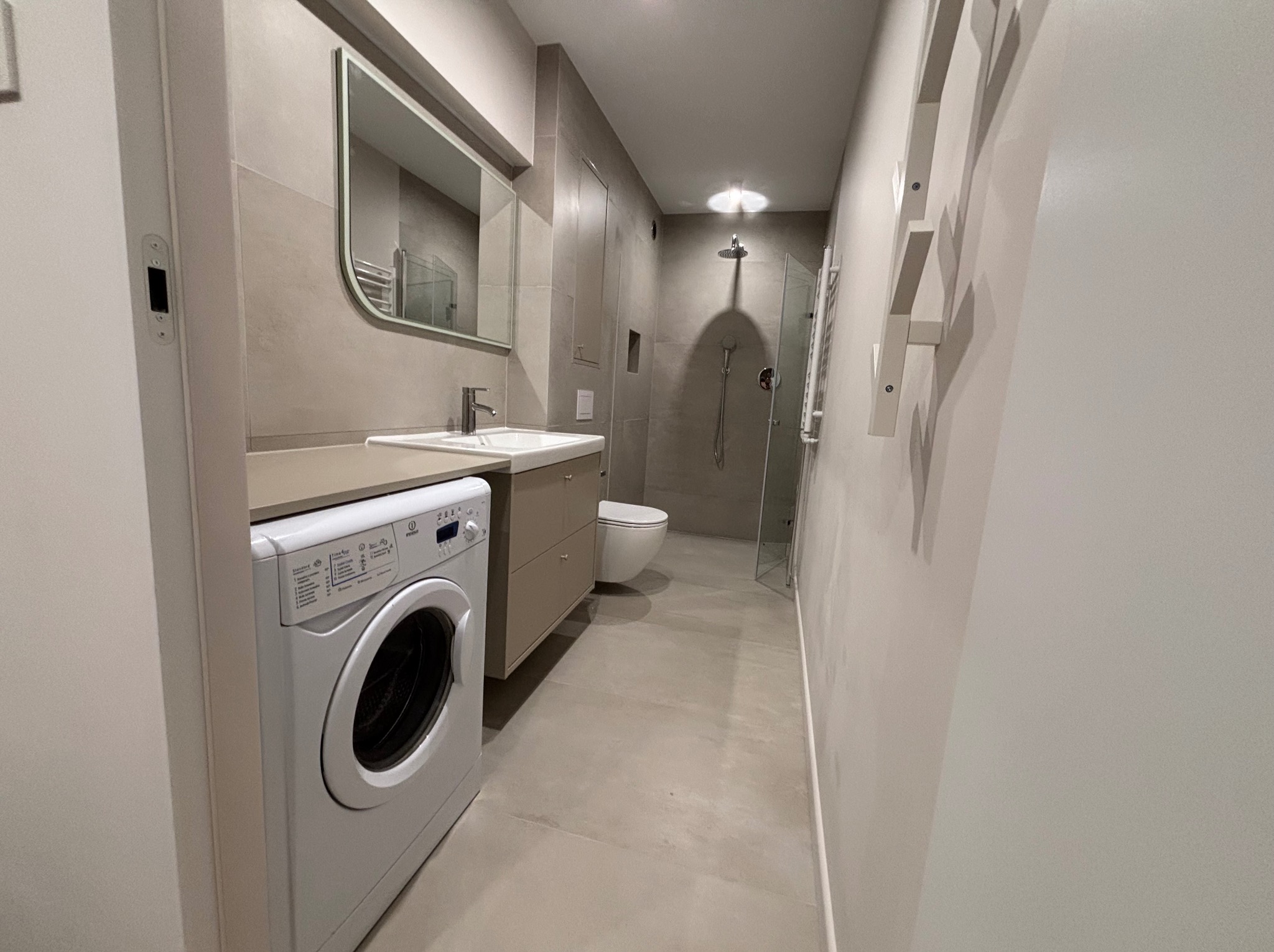 Jasna łazienka po remoncie: pralka, umywalka z szafką, lustro w zaokrąglonej ramie, kabina prysznicowa ze szklaną ścianką i toaleta w tle. Minimalistyczny design.