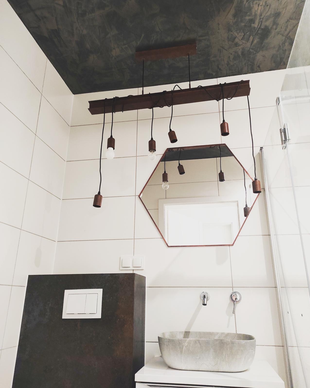 Nowoczesna łazienka z heksagonalnym lustrem, betonową umywalką i lampą z żarówkami Edisona. Ciemny sufit kontrastuje z białymi płytkami.