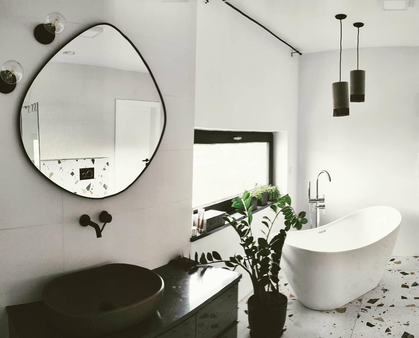 Nowoczesna łazienka z owalnym lustrem, czarną armaturą i wolnostojącą wanną na tle mozaikowej podłogi. Minimalistyczne oświetlenie i roślina doniczkowa dodają elegancji.