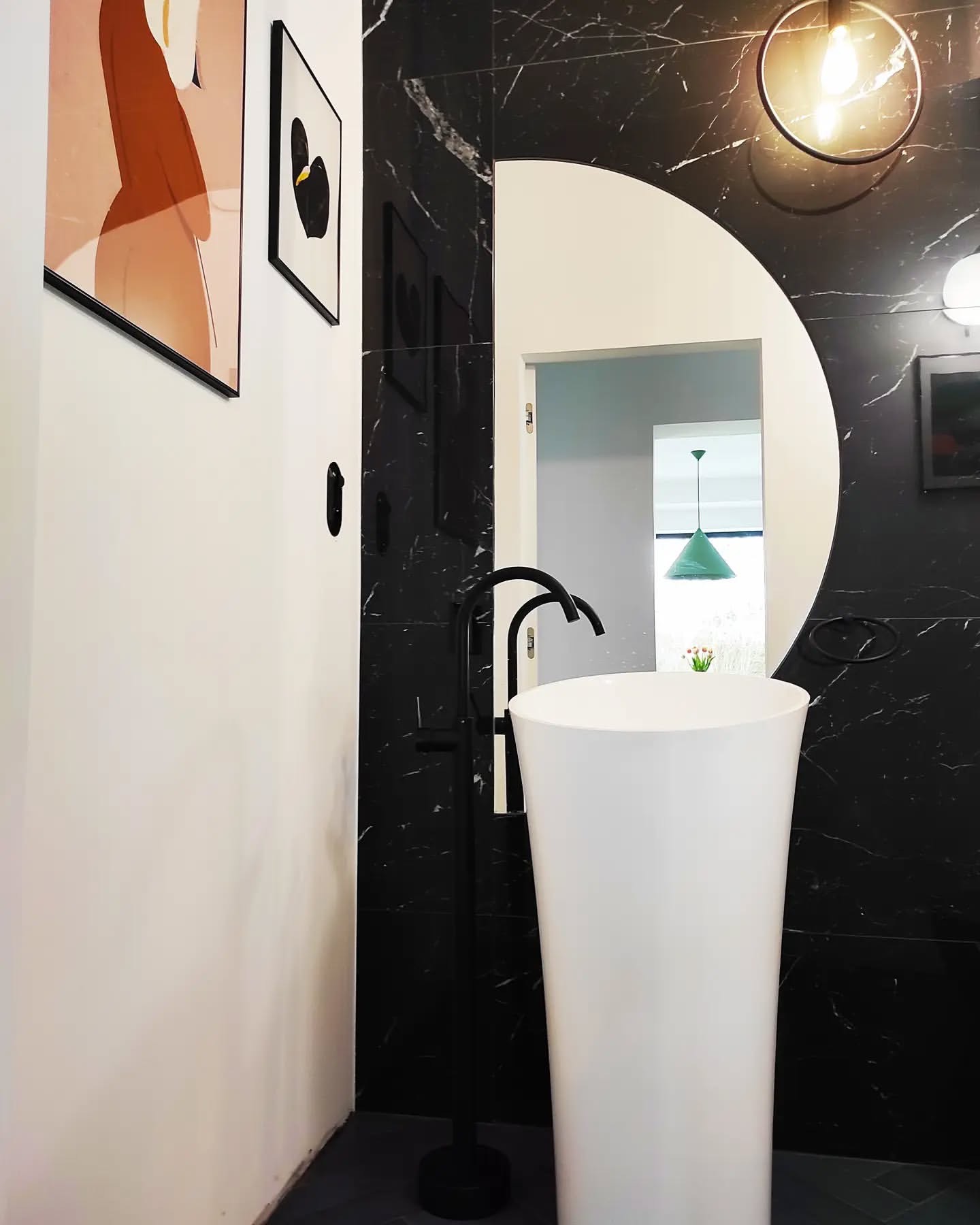 Nowoczesna łazienka z białą umywalką wolnostojącą i czarną armaturą na tle marmurowych płytek. W tle widoczne lustro z okrągłym wycięciem i dekoracje ścienne.