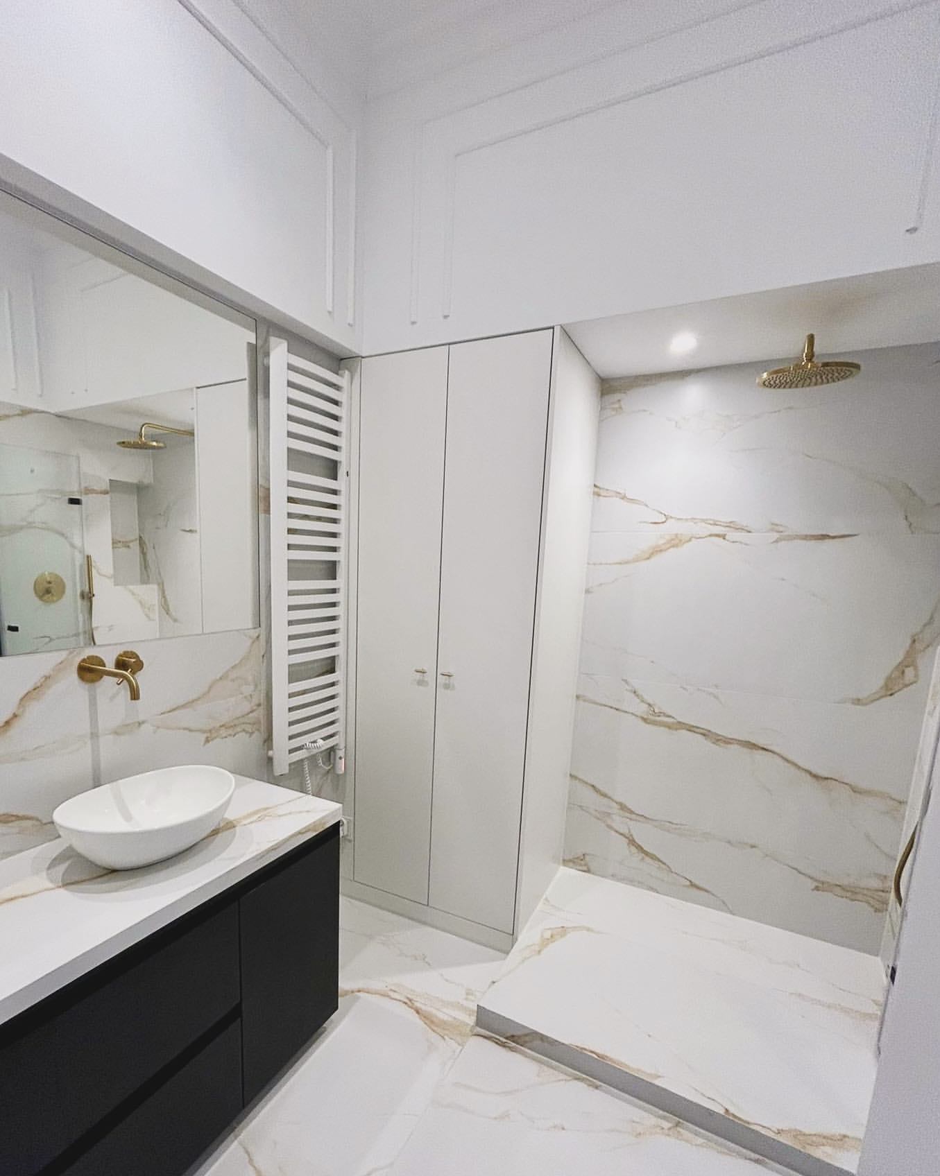 Elegancka łazienka po remoncie: białe marmurowe płytki z złotymi żyłami, czarna szafka pod umywalką, złota armatura, kabina prysznicowa typu walk-in i biały grzejnik.