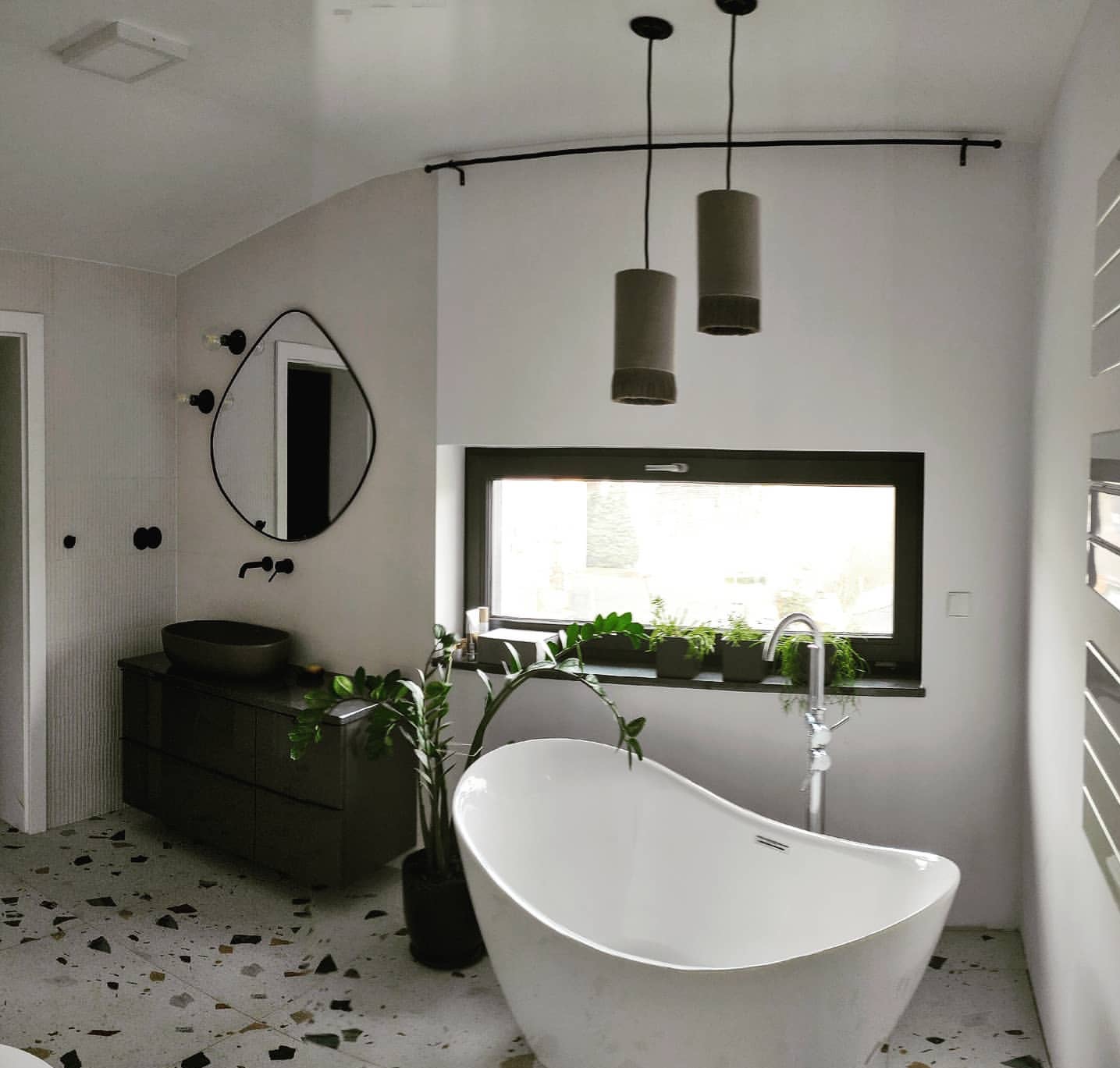 Nowoczesna łazienka z wolnostojącą wanną, umywalką nablatową i lustrem o nieregularnym kształcie. Na podłodze lastryko, a nad wanną wiszą lampy cylindryczne.