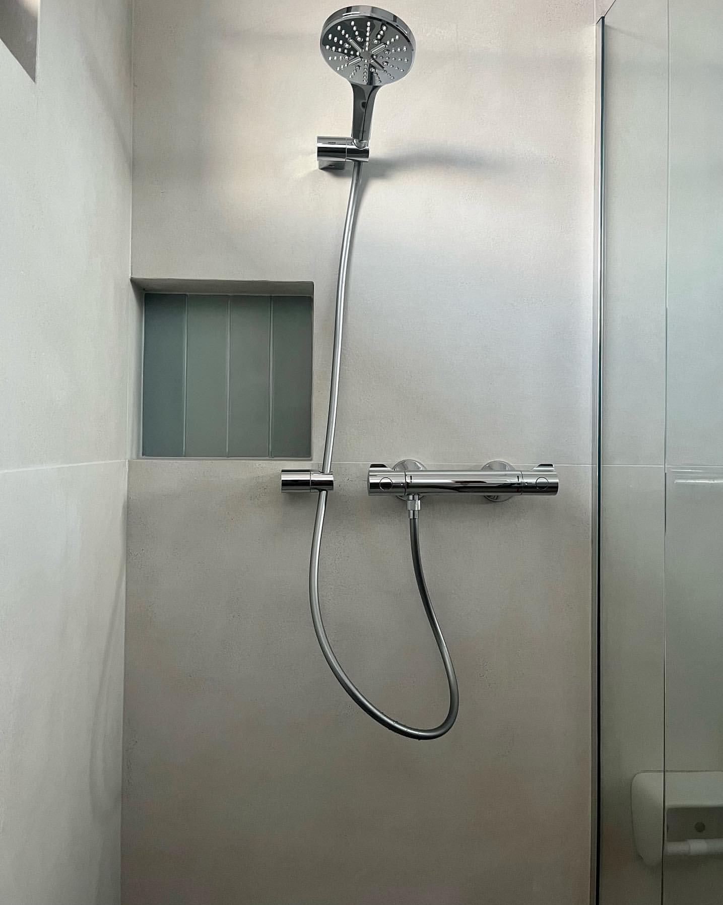 Nowoczesna kabina prysznicowa z deszczownicą i wnęką z szarymi płytkami. Minimalistyczny design, jasne ściany, chromowana armatura. Widok na szklane drzwi kabiny.