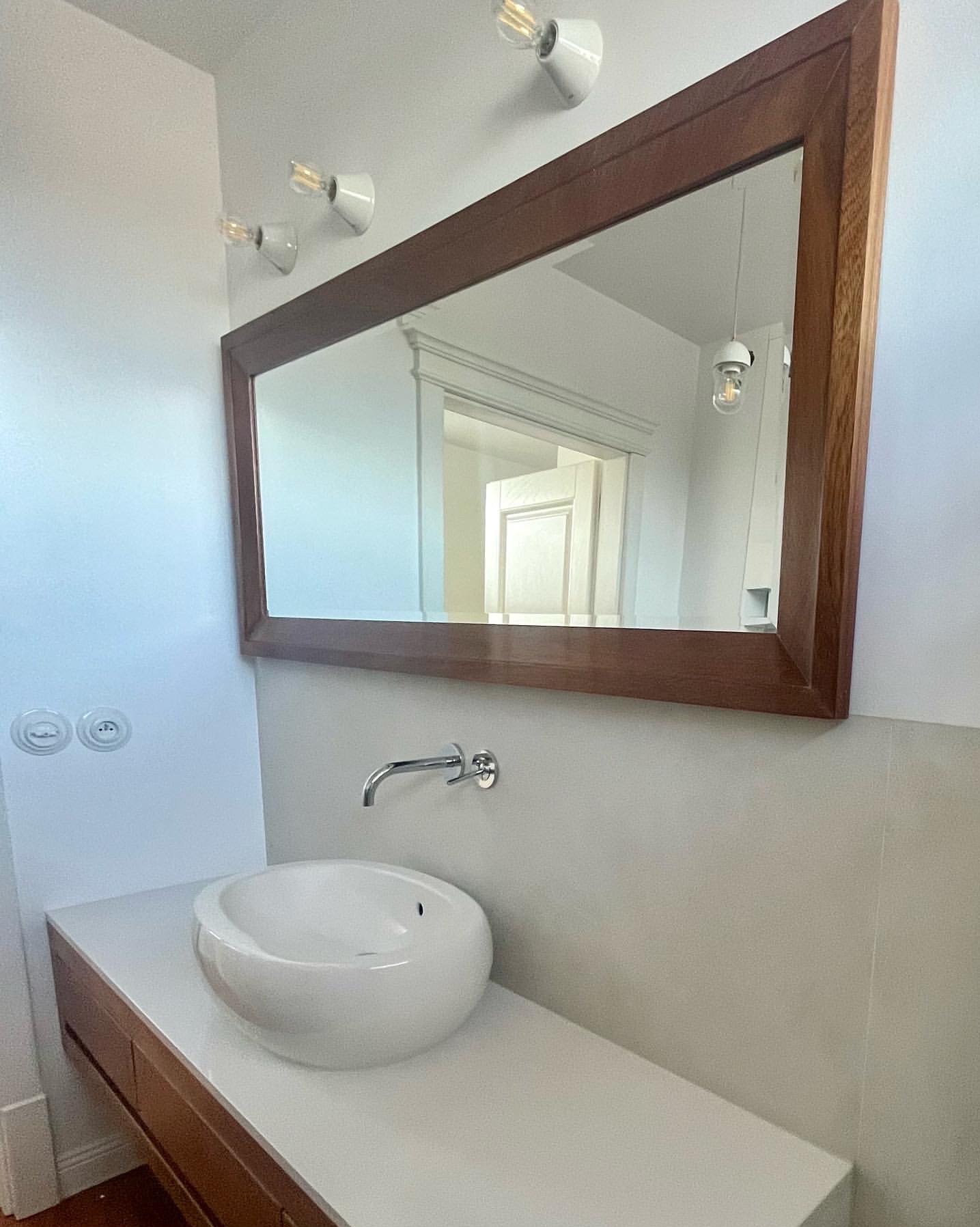 Nowoczesna łazienka z okrągłą umywalką, drewnianą szafką i dużym lustrem w drewnianej ramie. Minimalistyczne oświetlenie i jasne płytki tworzą elegancki wystrój.