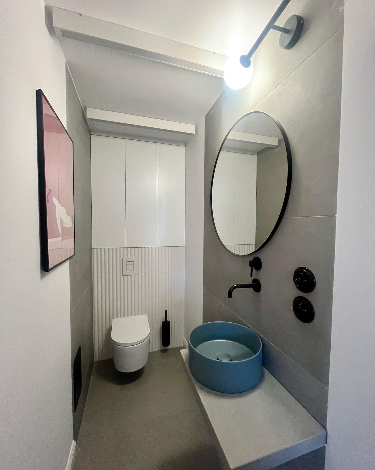 Nowoczesna łazienka w szarościach z akcentem błękitnej umywalki. Minimalistyczne oświetlenie, owalne lustro i geometryczne płytki tworzą spójną całość.