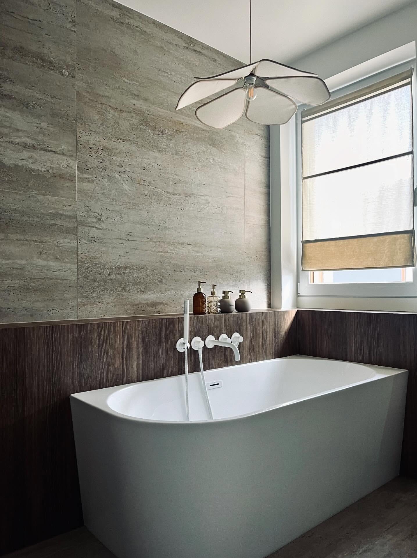 Nowoczesna łazienka z wolnostojącą wanną, armaturą w kolorze bieli i beżową ścianą z efektem trawertynu. Nad wanną designerska lampa w kształcie kwiatu.