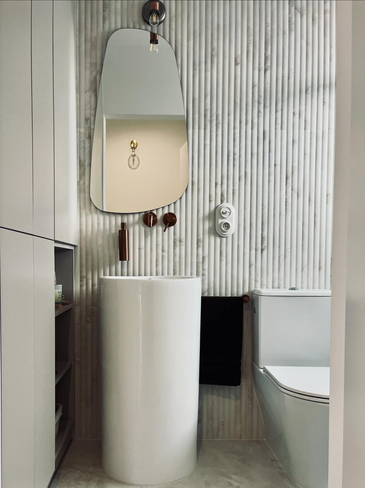 Nowoczesna łazienka z umywalką kolumnową, lustrem o nieregularnym kształcie i płytkami imitującymi marmur. Minimalistyczny design z akcentami w kolorze miedzi.