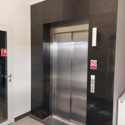 PPHU Dana,Danuta Cacak - Wykończenie windy kamieniem w ciemnym odcieniu, z widocznymi płytkami na ścianach i podłodze. Obok drzwi z oznaczeniem przeciwpożarowym.