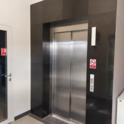 Wykończenie windy kamieniem w ciemnym odcieniu, z widocznymi płytkami na ścianach i podłodze. Obok drzwi z oznaczeniem przeciwpożarowym.