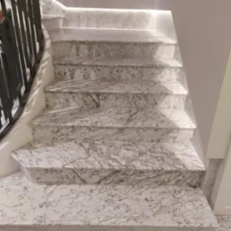 Marmurowe schody wewnętrzne z czarną, metalową balustradą i subtelnym oświetleniem LED pod stopniami, ukazujące eleganckie wykończenie.