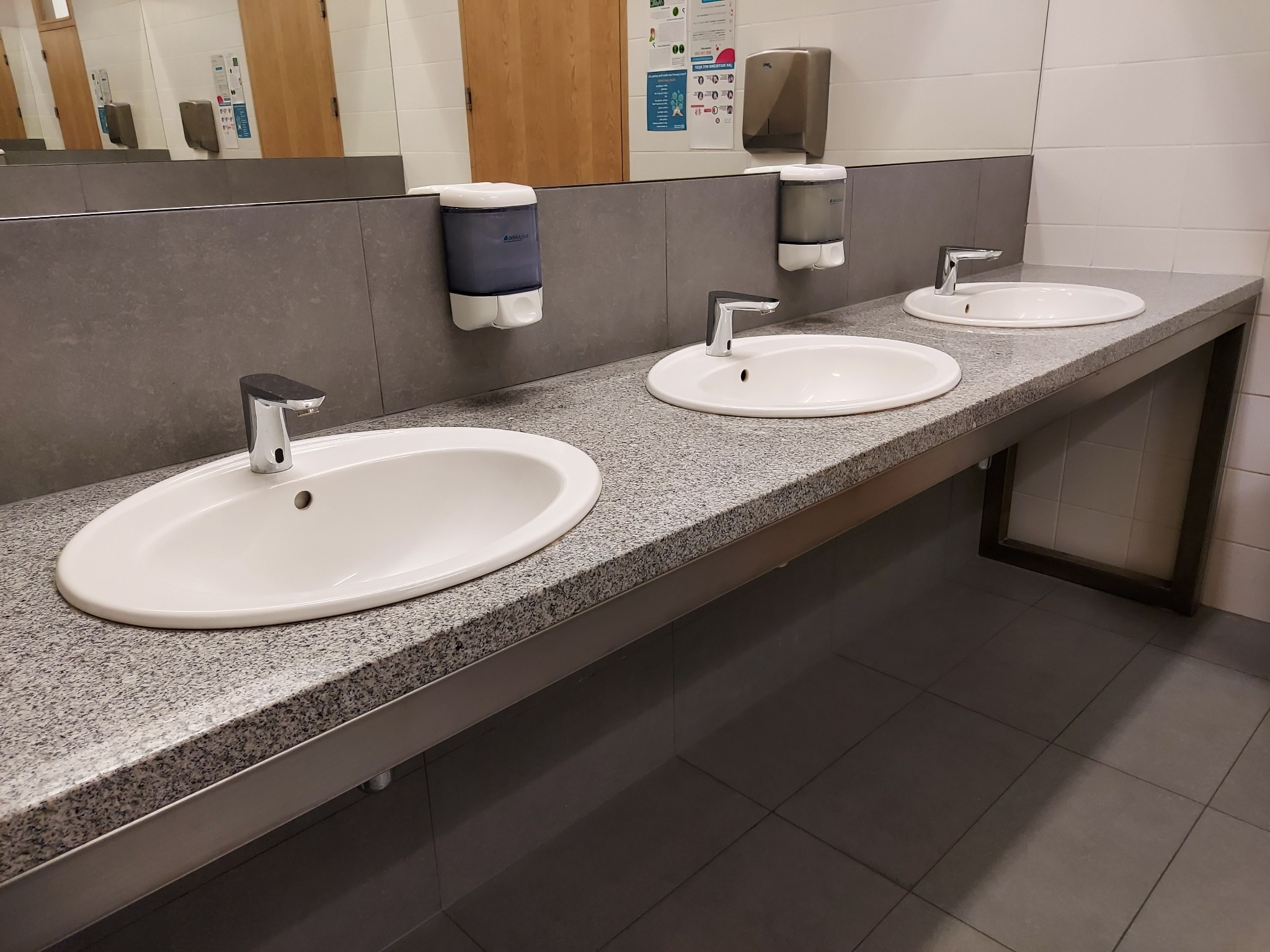 Blat łazienkowy z szarego kamienia z trzema umywalkami, dozownikami mydła i bateriami. Ujęcie z perspektywy lekko z dołu, widoczny fragment lustra i płytek na ścianie.