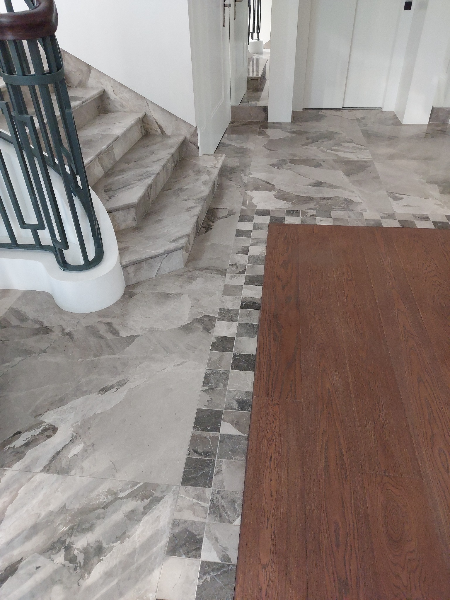 Marmurowe schody z metalową balustradą i podłoga z szarego marmuru z dekoracyjnym obramowaniem oraz panelami z ciemnego drewna w eleganckim wnętrzu.
