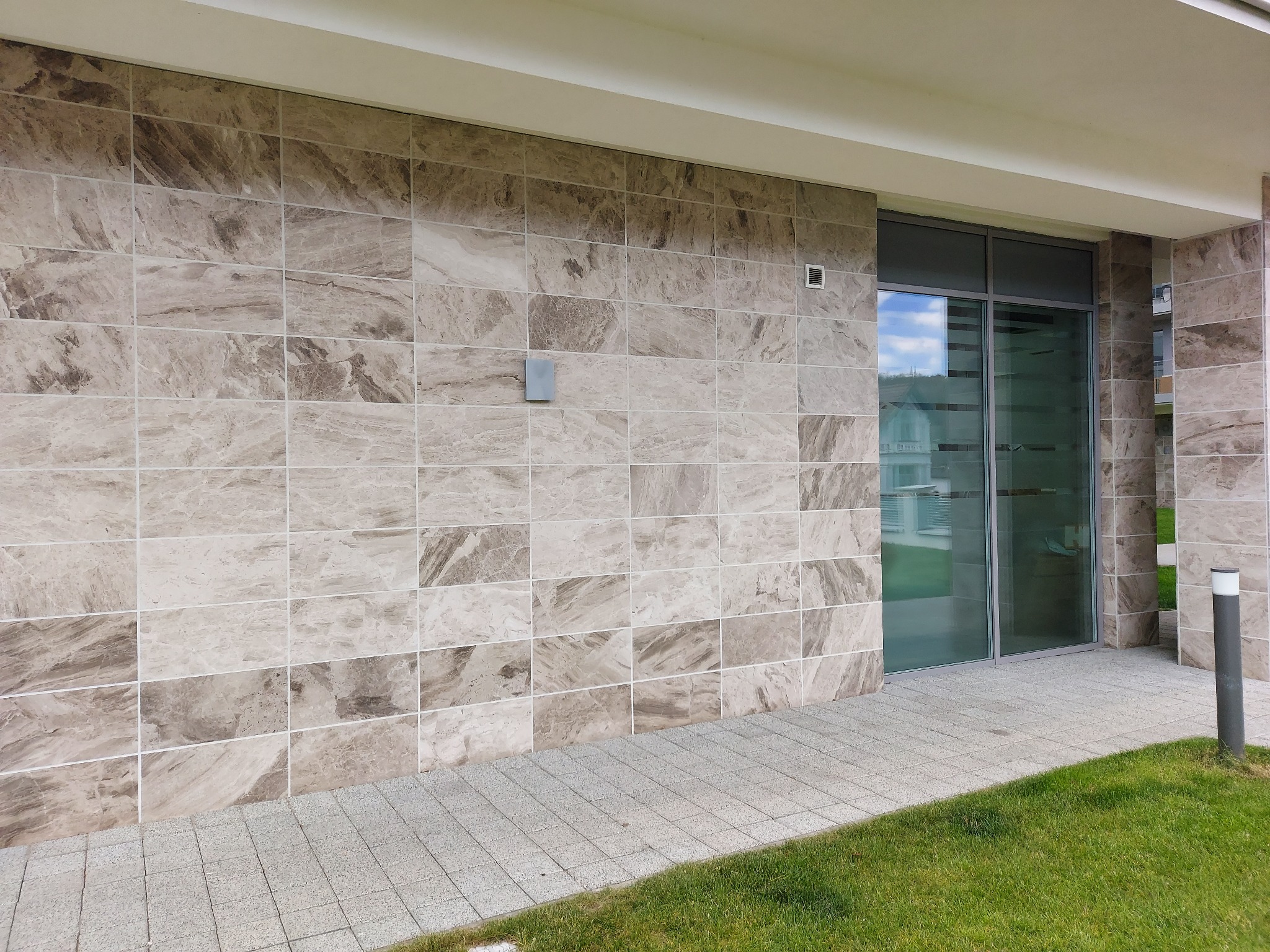 Elewacja budynku wykończona płytkami kamiennymi w odcieniach beżu i brązu, z widocznym fragmentem okna i chodnika z kostki brukowej. Detal architektoniczny.