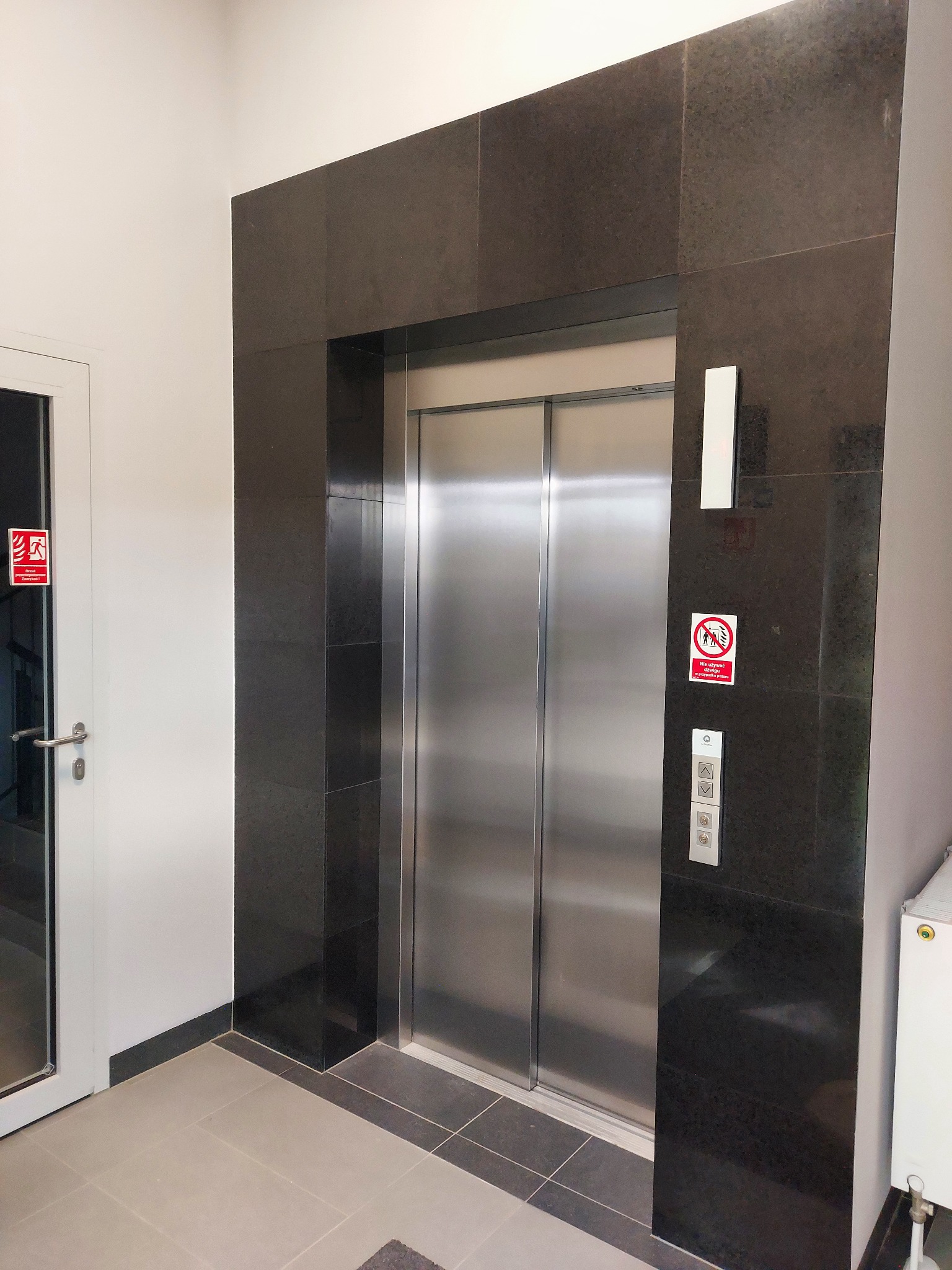 Wykończenie windy kamieniem w ciemnym odcieniu, z widocznymi płytkami na ścianach i podłodze. Obok drzwi z oznaczeniem przeciwpożarowym.