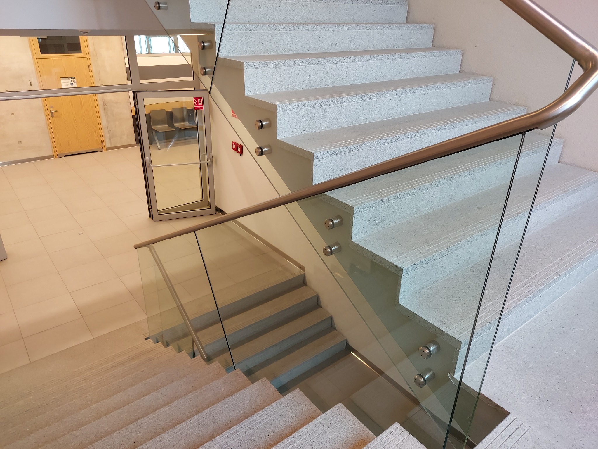 Nowoczesne schody kamienne z jasnego, drobnoziarnistego kamienia, z szklaną balustradą i metalową poręczą. Widok z góry, z widocznym fragmentem korytarza.