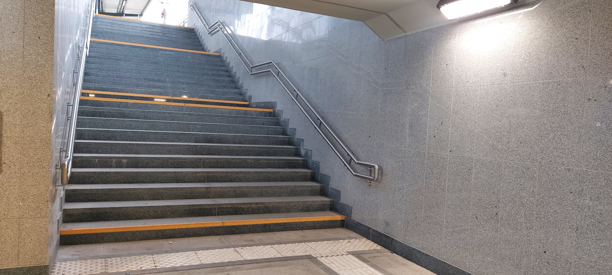 Kamienne schody z poręczą w Gorzowie Wielkopolskim, wykończone płytami o szarej, nakrapianej fakturze. Żółte pasy antypoślizgowe na krawędziach stopni.