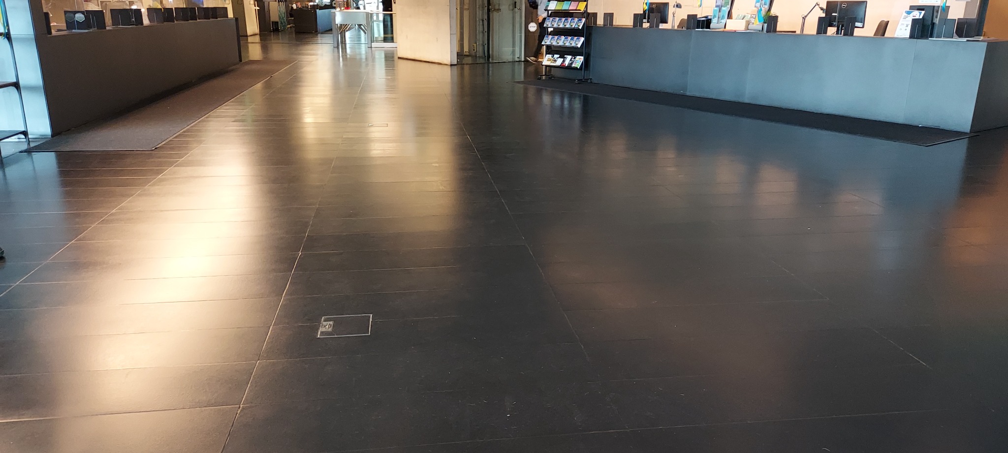 Błyszcząca, ciemna kamienna posadzka w dużej przestrzeni biurowej. Widoczne regularne spoiny i odbicia światła. Minimalistyczny design z elementami wyposażenia biura w tle.