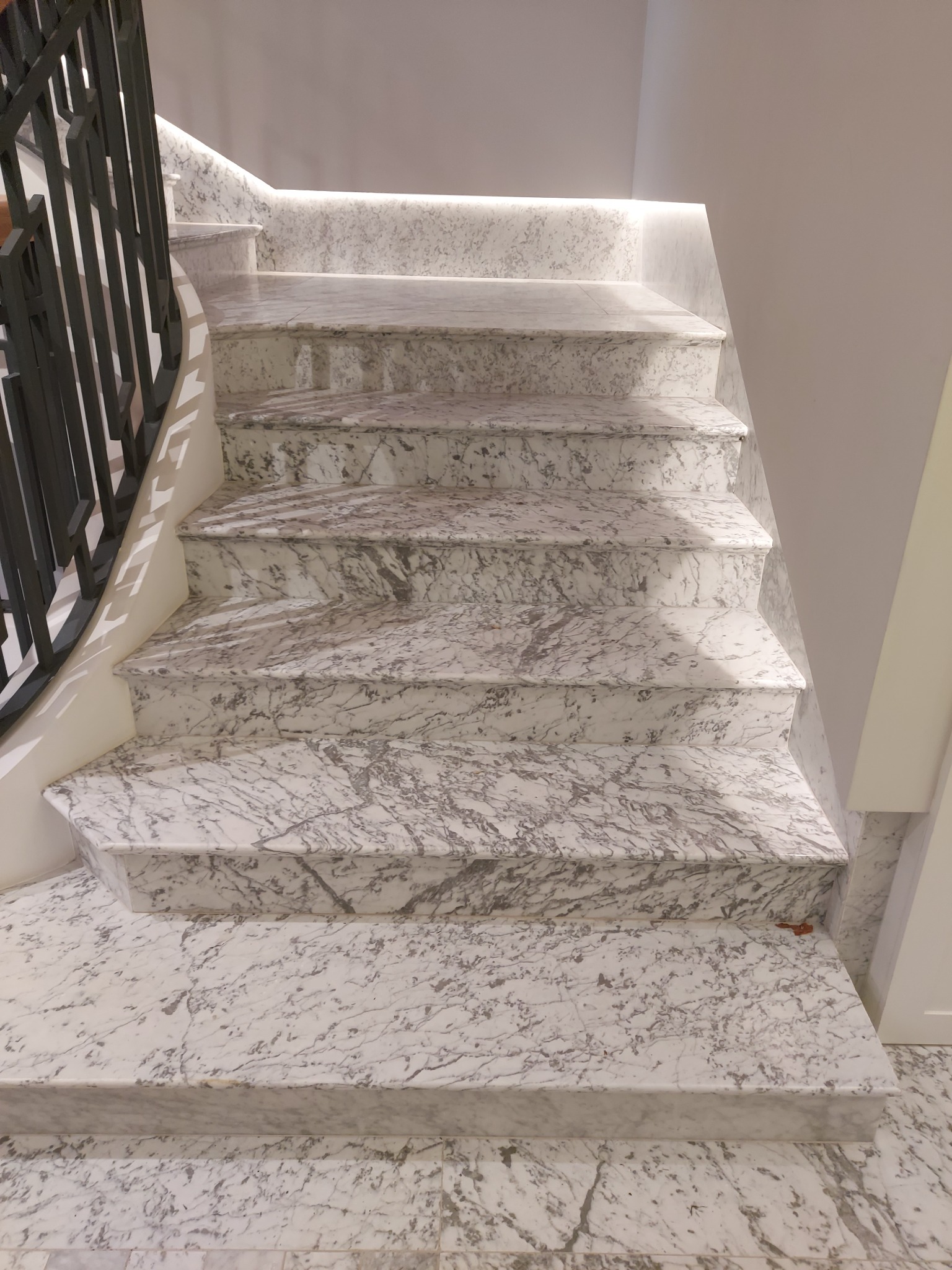 Marmurowe schody wewnętrzne z czarną, metalową balustradą i subtelnym oświetleniem LED pod stopniami, ukazujące eleganckie wykończenie.