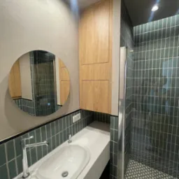 Nowoczesna łazienka z okrągłym lustrem, drewnianą szafką i kabiną prysznicową wyłożoną pionowymi, zielonymi płytkami. Podłoga w geometryczny wzór.