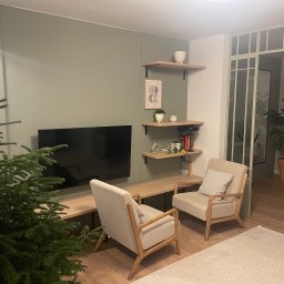 Sowiński - Wykończenia Wnętrz Mariusz Sowiński - Nowoczesny salon z zieloną ścianą, drewnianymi półkami i telewizorem. Dwa beżowe fotele i choinka tworzą przytulną atmosferę. Widoczne detale wykończenia wnętrza.