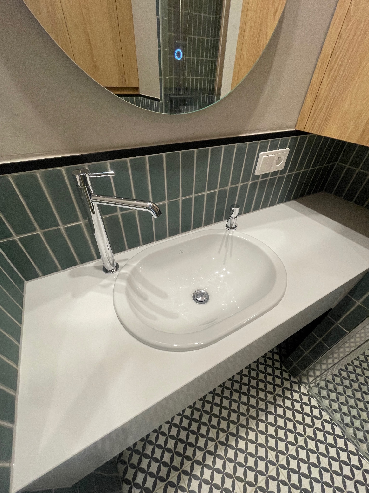 Biała umywalka z chromowaną baterią na blacie z zieloną, cegiełkową glazurą w tle i wzorzystą podłogą. Nowoczesna łazienka po remoncie.