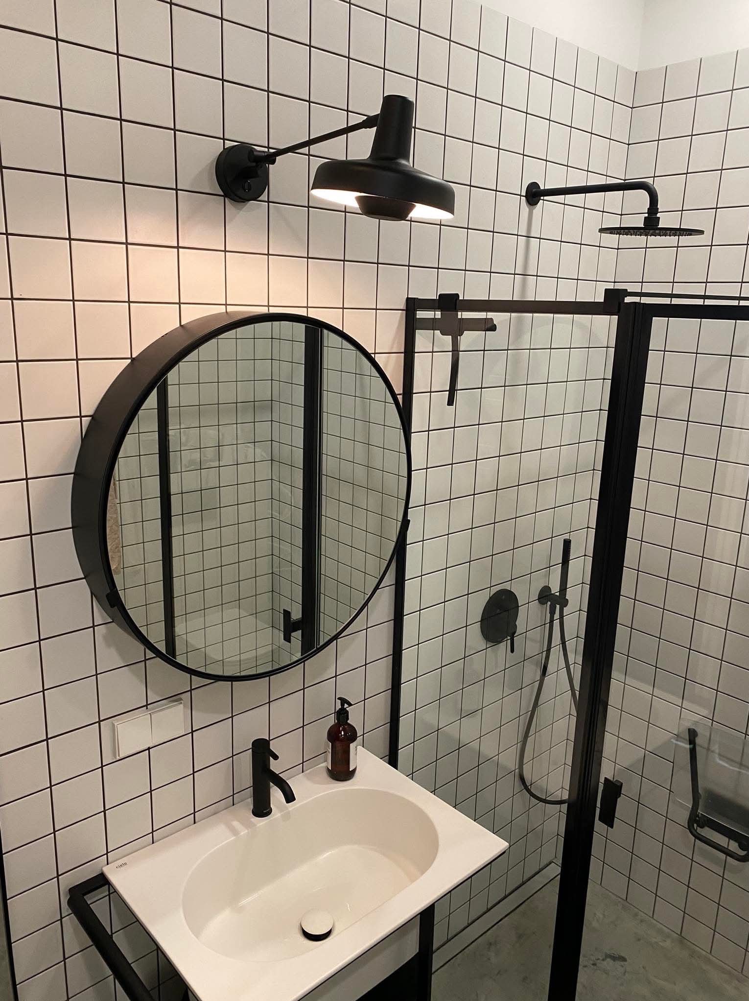 Nowoczesna łazienka z białymi kwadratowymi płytkami, czarną armaturą, okrągłym lustrem i kabiną prysznicową. Minimalistyczny design i oświetlenie punktowe.
